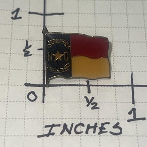May include: A metal lapel pin shaped like the North Carolina state flag, featuring a black vertical stripe with a gold star and the letters N and C, alongside red and yellow horizontal stripes. It measures approximately 0.75 inches in width.