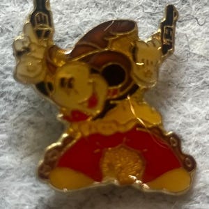 May include: A gold-colored pin featuring a cartoon character in a cowboy hat, holding two guns. The character has a yellow face, red clothing, and a brown hat. The pin has a detailed design with black accents.