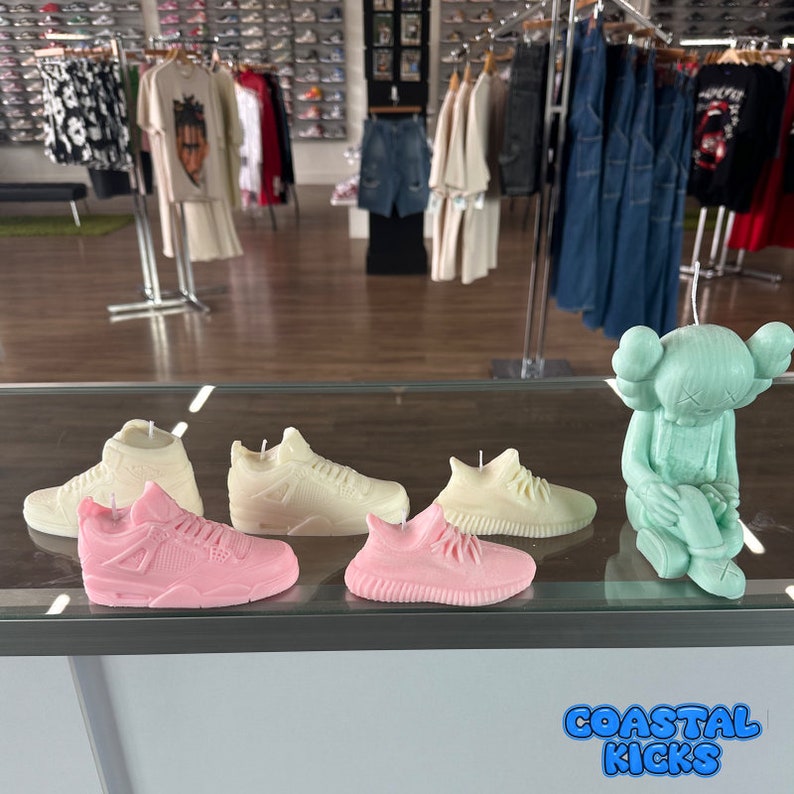 May include: A display of five sneaker-shaped candles, including a pair of pink Air Jordan 4s, a pair of white Yeezy 350s, and a pair of white Air Force 1s. A green, cartoon-like figure is also on display. The text "Coastal Kicks" is visible in the bottom right corner of the image.