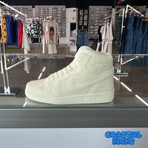 May include: A white high-top sneaker with a white swoosh logo and a white sole. The sneaker is on a glass display case with the text "Coastal Kicks" in blue letters on the bottom of the image.