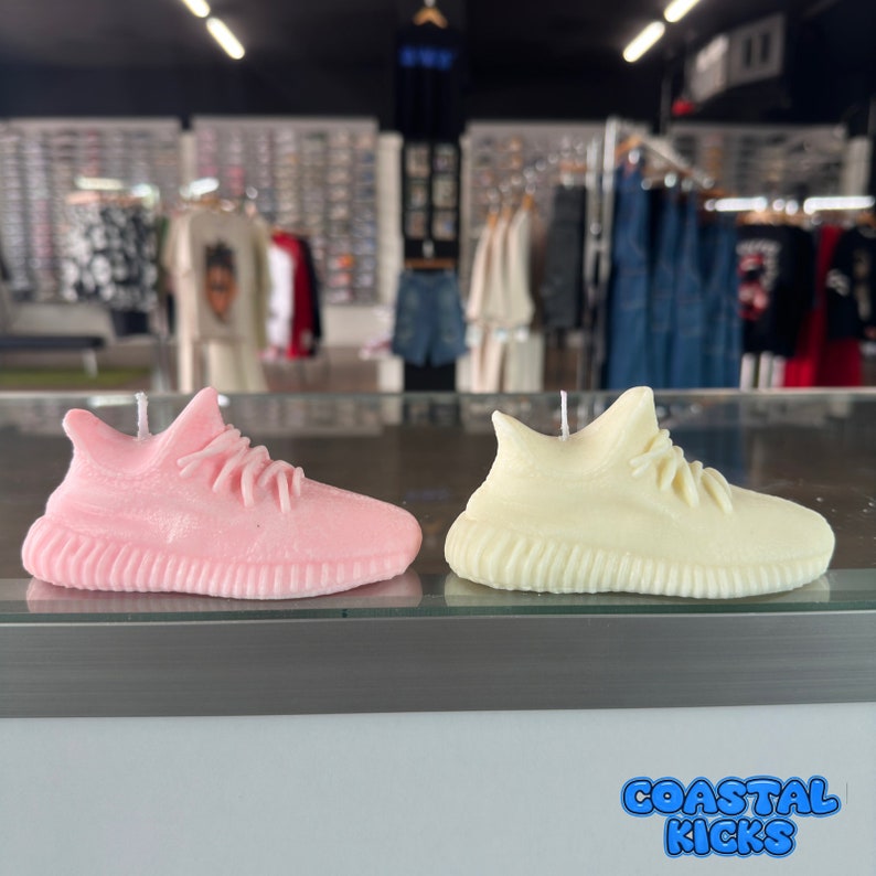 May include: Two sneaker-shaped candles, one pink and one cream, on a display shelf with the text "Coastal Kicks" in blue and white.