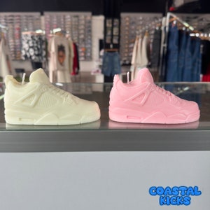May include: Two sneaker-shaped candles, one white and one pink, are displayed on a counter. The candles are detailed with the classic design of a popular athletic shoe. The text "Coastal Kicks" is visible in the bottom right corner of the image.