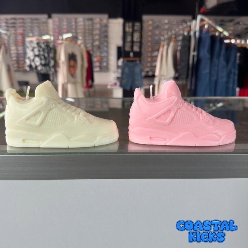 May include: Two sneaker-shaped candles, one white and one pink, are displayed on a counter. The candles are detailed with the classic design of a popular athletic shoe. The text "Coastal Kicks" is visible in the bottom right corner of the image.