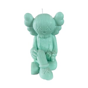May include: A green candle shaped like a sitting figure with large ears and a simple face. The candle has a textured surface and a small wick on top.