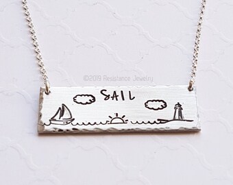 May include: A silver necklace with a rectangular pendant that says "SAIL" with a sailboat, sun, and lighthouse illustration. ©2019 Resistance Jewelry