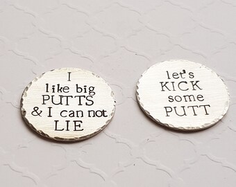 May include: Two silver golf ball markers with engraved sayings. The first marker says "I like big putts & I can not lie". The second marker says "Let's kick some putt".
