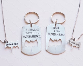 May include: Two silver keychains with a Batman silhouette cutout. One keychain says "Husband. Father. Superhero." and the other says "Dad is my Superhero." A silver necklace with a smaller Batman silhouette cutout that says "sidekick" is also pictured.