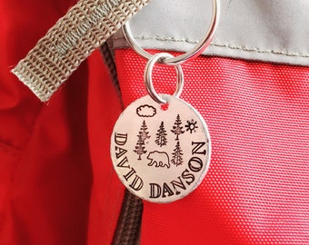 May include: A silver keychain with a round tag engraved with the name "DAVID DANSON" and a design of a bear, trees, a cloud, and a sun.