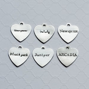 May include: Six silver heart-shaped charms with different words engraved on them. The words are "Stargazer", "Melody", "Newsprint", "Blackjack", "Juniper", and "ARCADIA".