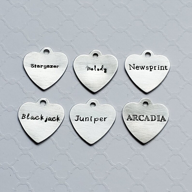 May include: Six silver heart-shaped charms with different words engraved on them. The words are "Stargazer", "Melody", "Newsprint", "Blackjack", "Juniper", and "ARCADIA".