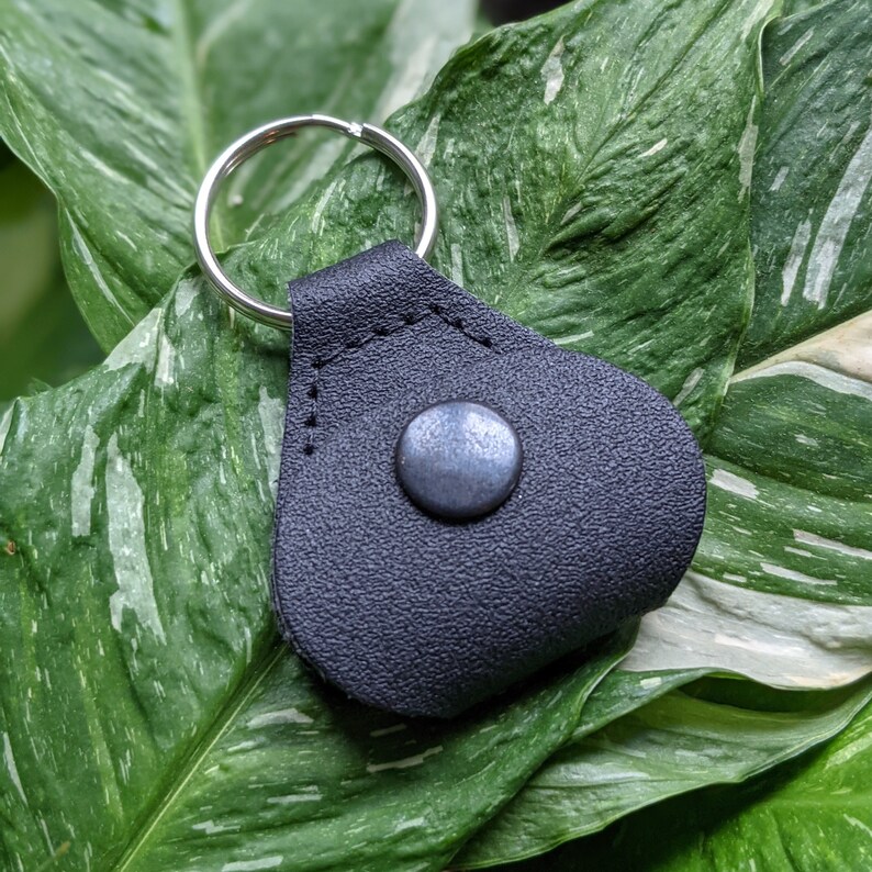 May include: A black leather key chain with a silver ring and a silver metal button. The keychain is on a green and white leaf background.
