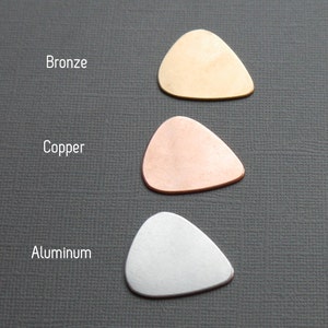 May include: Three guitar picks made of different metals: bronze, copper, and aluminum. The picks are arranged vertically, with the bronze pick at the top, the copper pick in the middle, and the aluminum pick at the bottom.