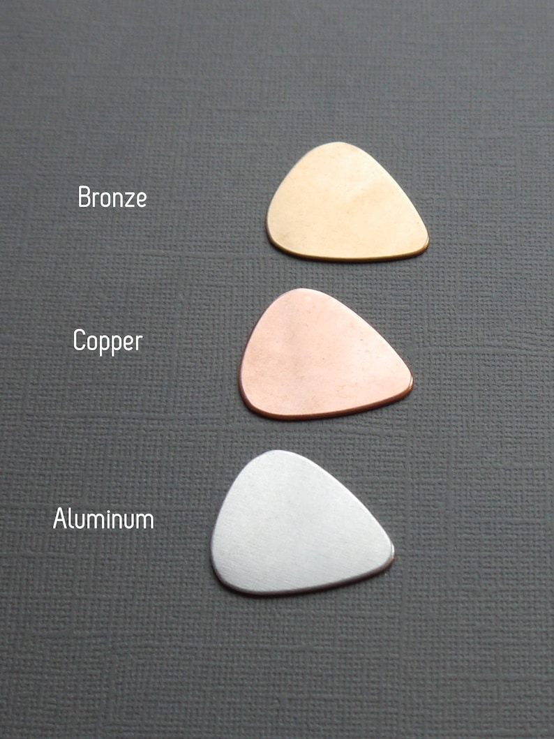 May include: Three guitar picks made of different metals: bronze, copper, and aluminum. The picks are arranged vertically, with the bronze pick at the top, the copper pick in the middle, and the aluminum pick at the bottom.