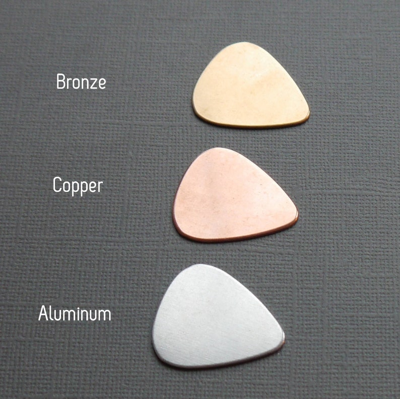 May include: Three guitar picks made of different metals: bronze, copper, and aluminum. The picks are shaped like a standard guitar pick and are arranged vertically from top to bottom.