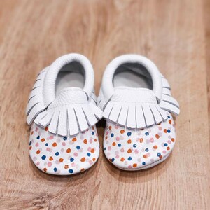 May include: White leather baby moccasins with a fringe detail and a colorful polka dot pattern in shades of pink, orange, blue, and white.