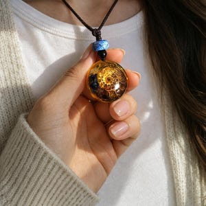 Könnte beinhalten: Eine Halskette mit Anhänger, die einen runden, bernsteinfarbenen Harzanhänger mit einem Spiralmuster aufweist. Der Anhänger hängt an einer schwarzen Kordel mit blauen und schwarzen Perlen. Die Halskette wird in einer Hand gehalten.