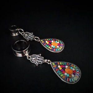 May include: A pair of silver-toned ear gauges with a dangling teardrop charm. The charms feature a silver Hamsa hand and a colorful mosaic of small, round gemstones in various colors, including orange, blue, green, and yellow.