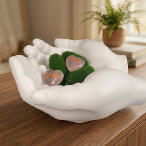 Peut inclure: Une sculpture en céramique blanche en forme de deux mains jointes tenant un amas de mousse verte et deux pierres roses translucides en forme de cœur. Chaque pierre rose porte le mot "love" imprimé en lettres orange sur sa surface.