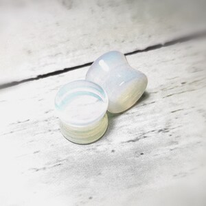 May include: Two white opal ear gauges, one is a flat disc and the other is a teardrop shape. The gauges are on a white wooden surface.