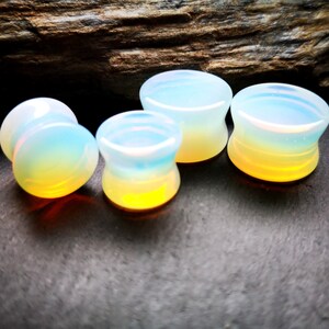 May include: Four iridescent opal ear gauges in various sizes. The gauges are a light blue color with a yellow-orange hue at the bottom.