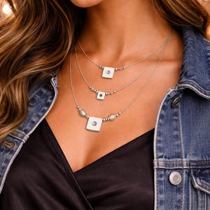 Può includere: Collana multistrato con tre catene d'argento. Ogni catena ha un ciondolo quadrato con una pietra trasparente centrale e piccole perline. La modella indossa un top nero e una giacca di jeans. Un gioiello delicato.