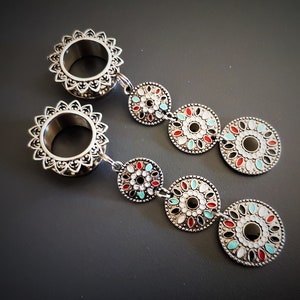 May include: A pair of silver ear gauges with dangling charms. Each charm is a silver circle with a black center and a white, red, and turquoise enamel design.