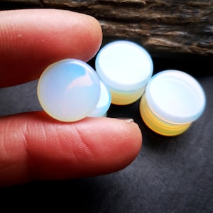 May include: Four iridescent opal plugs, two are held in a hand, and two are on a dark surface. The plugs are round and have a smooth, polished finish.