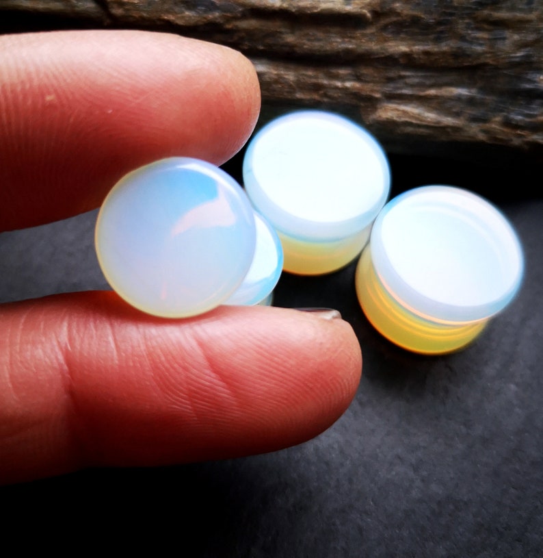 May include: Four iridescent opal plugs, two are held in a hand, and two are on a dark surface. The plugs are round and have a smooth, polished finish.