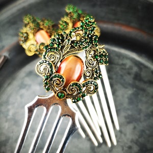 Könnte beinhalten: Zwei silberne Haarkämme mit verzierten goldenen und grünen Juwelenbesatz. Die Verzierungen zeigen einen großen, ovalen, orangefarbenen Edelstein.