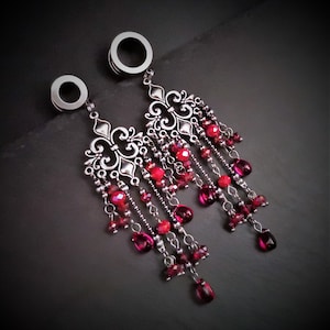 May include: A pair of silver ear gauges with dangling red gemstone charms. The charms are attached to a silver filigree design.