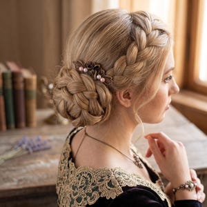 May include: A woman with blonde hair styled in a braided updo, adorned with a bronze hair comb featuring pink pearls. She wears a dark dress with gold lace detailing, a matching necklace, and a bracelet. The image has a vintage aesthetic.