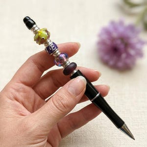 Könnte beinhalten: Ein schwarzer Tintenstift mit einer Reihe dekorativer Perlen am oberen Ende, darunter eine limettengrüne froschförmige Perle, eine violette Perle mit dem Buchstaben E, eine schimmernde irisierende Perle und eine einfarbige dunkelviolette Perle.