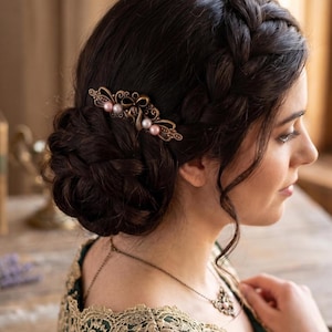 May include: A bronze-toned hair clip with a decorative bow and pearl accents. The clip is set in dark hair styled in an updo. The woman is wearing a green and gold lace top and a matching necklace.
