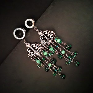 May include: A pair of silver ear gauges with dangling green crystal charms. The charms are shaped like a fleur-de-lis and have multiple strands of beads.