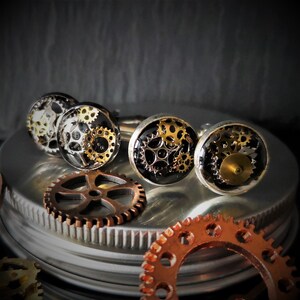 May include: Steampunk cufflinks featuring detailed gear designs in silver, gold, and bronze. The cufflinks are set against a dark backdrop, with loose gears scattered around, creating a vintage, industrial aesthetic.