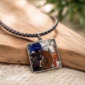 May include: A square pendant necklace with a silver frame and a black leather cord. The pendant is filled with a variety of colorful stones and a spiral shell. The stones include blue, white, red, and brown.