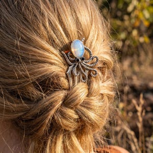 Könnte beinhalten: Ein silberfarbener Haarkamm mit einem Oktopus-Design. Der Kamm hat einen großen, ovalen, irisierenden Stein in der Mitte, mit schwarzen Steinen und Tentakeln. Der Kamm ist in einen blonden Zopf gesteckt.
