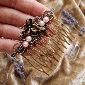 May include: An antique bronze hair comb with an ornate design featuring a bow and decorative swirls. The comb is adorned with faux pearls in white and pink. The comb's tines are long and slender, and the overall style is vintage.