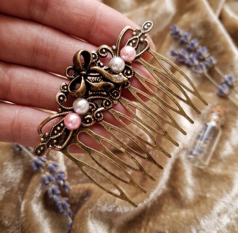 May include: An antique bronze hair comb with an ornate design featuring a bow and decorative swirls. The comb is adorned with faux pearls in white and pink. The comb's tines are long and slender, and the overall style is vintage.