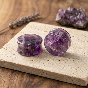 Könnte beinhalten: Zwei kreisförmige Ohrplugs aus poliertem violettem Amethyststein. Die Plugs haben eine glatte, glänzende Oberfläche mit natürlichen kristallinen Einschlüssen, die im tiefvioletten Material sichtbar sind und die Muster des Edelsteins zeigen.