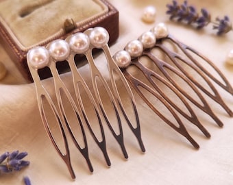 May include: Two hair combs with pearl accents. One comb is silver-toned, the other is bronze-toned. Each comb has multiple tines and a row of pearls. The combs are displayed on a cream-colored surface with a brown jewelry box in the background.