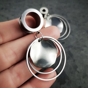 Könnte beinhalten: Silberne Edelstahl-Ohrmanschetten mit einer großen runden Scheibe und zwei kleineren Ringen, die unten hängen.