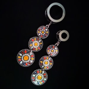 May include: A pair of silver ear gauges with dangling charms. The charms are round and feature a colorful floral design with red, yellow, white, and blue enamel.