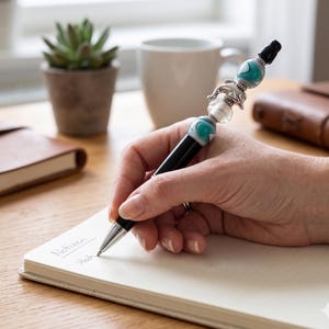 Könnte beinhalten: Ein schwarzer Kugelschreiber mit einem dekorativen Oberteil, das mit zwei türkis-weißen Wirbelperlen, einer transparenten Perle und einem silberfarbenen Delfin-Anhänger verziert ist. Der Stift hat eine silberne Spitze und eine schwarze Kappe am Ende.