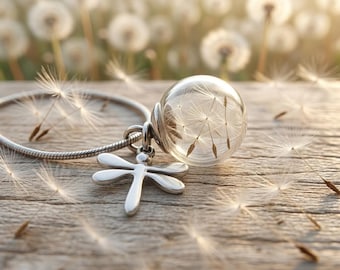 May include: A silver bracelet with a dragonfly charm and a clear glass orb pendant containing dandelion seeds. The jewelry rests on a wooden surface, with a blurred background of dandelions.
