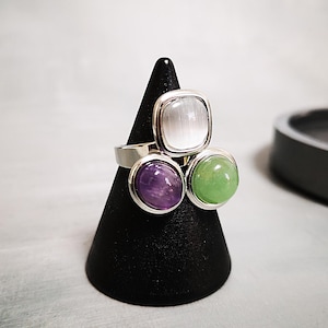 Könnte beinhalten: Ein silberner Ring mit drei Edelsteinen: ein quadratischer, weißer Stein und zwei runde Steine, einer lila und einer grün. Der Ring wird auf einem schwarzen, kegelförmigen Ständer präsentiert.