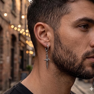 May include: A silver dagger earring with a hoop closure. The dagger has intricate detailing on the hilt and a pointed blade. The earring is worn on the ear of a person with a beard.