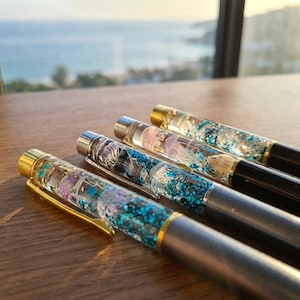 May include: Four pens with clear barrels filled with liquid, blue glitter, and small decorative elements including jellyfish figures and seashells. Each pen features a metallic clip and a solid coloured base in silver or black with gold accents.