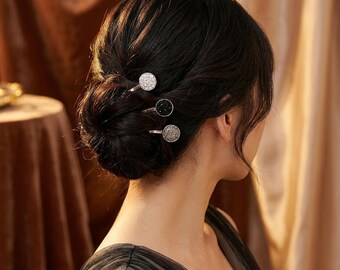 May include: Three decorative hair pins are inserted into a bun hairstyle. The pins have round, embellished heads in silver, black, and crystal. The woman is wearing a black dress, and the background is a warm, neutral color.