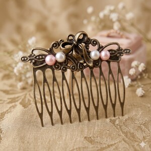 May include: An antique bronze hair comb with a decorative bow and three faux pearls in pink and white. The comb has multiple tines and is designed for securing hair. The comb is likely for a wedding or special occasion.
